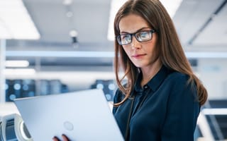 Dark-haired woman wearing glasses looks intently at a laptop. The screen reflects in her glasses.